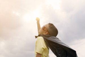 A young boy wearing a cape reaching his hand to the sky in bright daylight evoking a sense of wonder